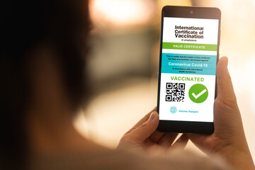 Smartphone displaying a valid digital vaccination certificate passport for COVID-19 in female's hand, public area background. Vaccination, disease immunity passport, health and safty travel concepts