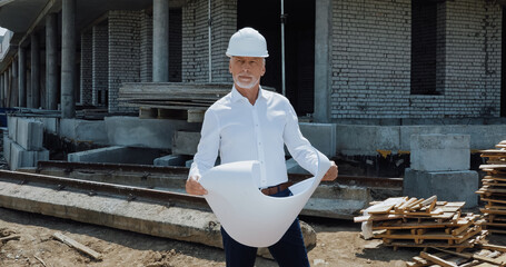 mature engineer in hard hat holding blueprint on construction site