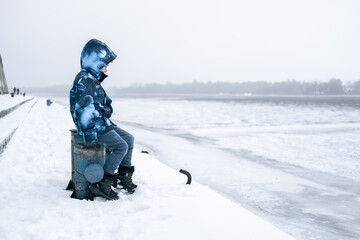 Fototapeta na wymiar A young boy sits in the winter sadly near the frozen river.