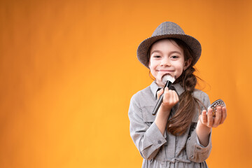 Adorable girl in a gray hat in a pigtail paints her face with a brush holding a small mirror in her hand