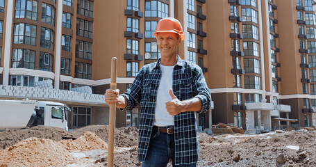 Fototapeta premium Smiling builder holding shovel and showing thumb up on construction site