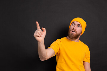Portrait of bearded european man in yellow hat on black background shouts angrily, reproachfully accuses someone, points directly to left, blames someone