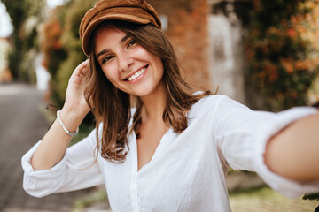 Young woman with short hair wearing white cotton blouse and brown hat. Girl poses outdoors and...