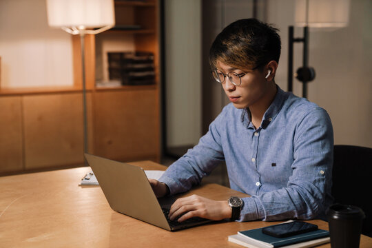 Focused Asian Guy Working With Laptop While Sitting At Table In Office