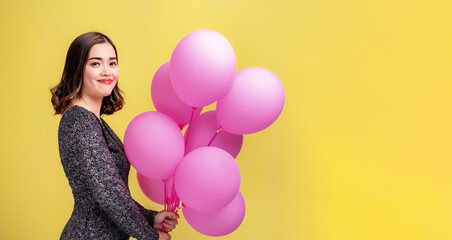 Beautiful brown haired Asian woman smiling and holding pink balloons on yellow background with copy space on the right.