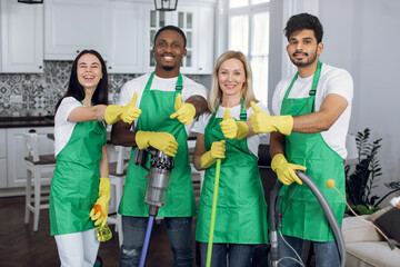 Smiling multiracial housekeepers in green aprons and yellow gloves showing thumbs up at work. Happy professional cleaner team gesturing on camera at bright room.