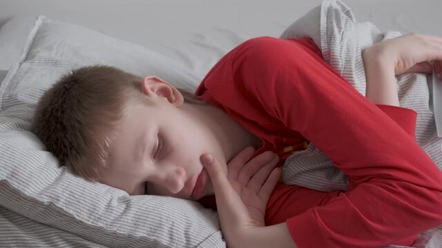 Sick teenage boy in red pajamas lies on his side in bed and coughs. Sick look, chapped lips
