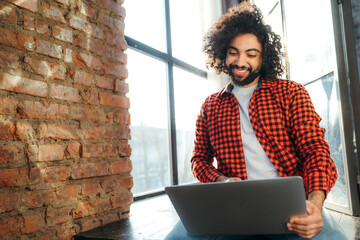 Stylish egyptian student in red plaid shirt sitting on floor smiling and looking at laptop