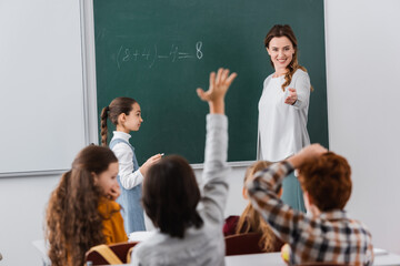 smiling teacher standing and chalkboard and pointing at pupils on blurred foreground