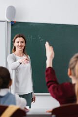 happy teacher pointing at schoolgirl raising hand on blurred foreground