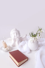 still life in high key style, old paper book in a dark red binding on white table, candle, bouquet of half snowdrops in vase, concept of changing seasons, reading literature