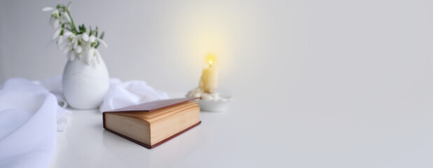 panorama, banner of still life in high key style, old paper book in a dark red binding on white table, candle, bouquet of half snowdrops in vase, concept of changing seasons, reading literature