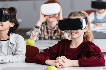 cheerful schoolkids gaming in vr headsets in classroom, blurred background