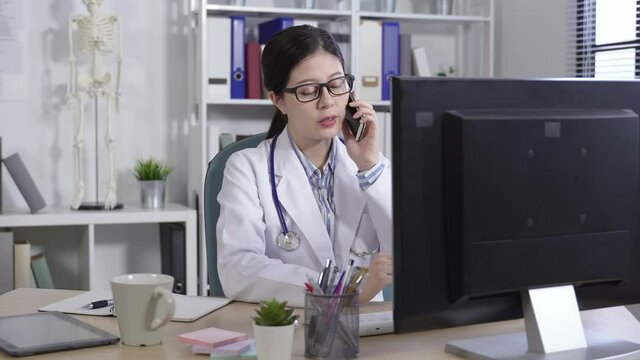 cheerful asian doctor seated at a office desk is checking on the computer while having a phone talk at a modern personal workplace.