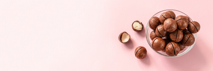 Raw macadamia nuts in a transparent glass bowl and scattered on a pink solid background. Minimal composition.