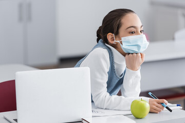 Schoolgirl in medical mask holding pens near apple and laptop on desk