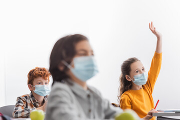 Schoolgirl in medical mask raising hand near classmates in class