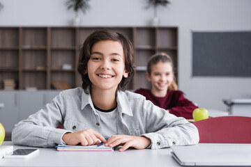 Smiling child looking at camera near devices and notebook on desk in classroom