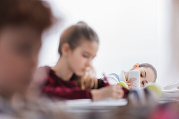 Obraz na płótnie Canvas Schoolchild using smartphone near classmates on blurred foreground in class