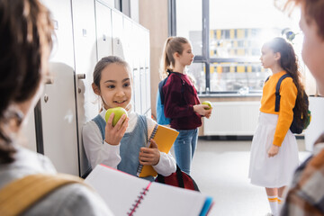 Happy child holding ripe apple and notebook while looking at schoolkids on blurred foreground