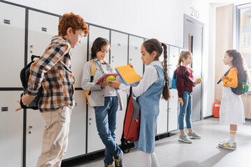Schoolgirl holding apple near boys with backpacks in school