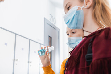 Schoolchild holding disinfector near friend in medical mask on blurred foreground