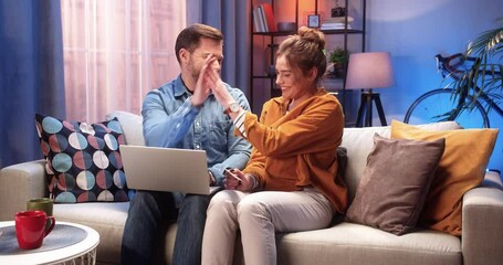 Happy young Caucasian married couple man and woman buying on internet on laptop making payment with credit card sitting on couch in house at night in good mood with satisfied faces, online shopping