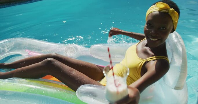 Attractive african american woman sitting on inflatable holding drink and smiling in swimming pool