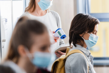 Teacher holding infrared thermometer and notebook near pupils in medical masks on blurred foreground