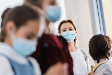 Teacher looking at schoolkids in medical masks on blurred foreground
