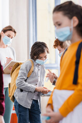 Schoolboys in medical masks cleaning hands with sanitizer near teacher on blurred background