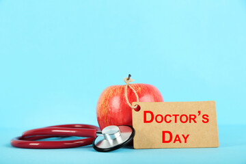 Text Doctor's Day with red apple and stethoscope on blue background