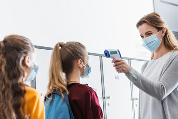 Teacher in protective mask using infrared thermometer on schoolgirls during quarantine in school