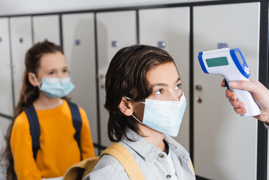 Teacher with infrared thermometer checking temperature of boy in protective mask in hall
