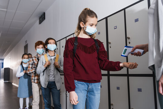 Teacher measuring temperature of schoolgirl in medical mask with infrared thermometer in hall
