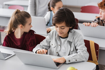 schoolboy typing on laptop in classroom near classmates on blurred background