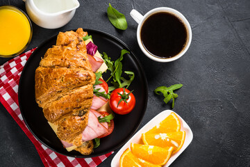 Croissant sandwich with ham, cheese and vegetables on wooden cutting board with cup of coffeee at black stone table. Top view with copy space.