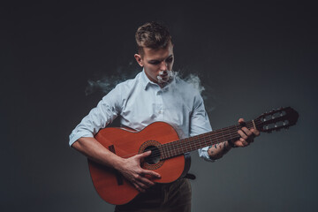 Portrait of a hipster guy dressed in formal clothing playing guitar in dark background and smoking cigarette.