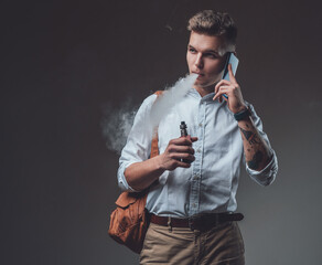 Isolated in dark background man. Handsome guy with stylish hairstyle talks on his smartphone smoking electric cigarette.