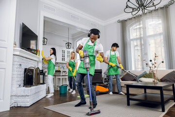 Handsome young african worker of cleaning service, cleaning up carpet and dancing, listening to music in headphones, while another members of cleaning team working on background.