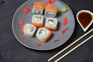 Rolls with salmon, rice, cheese, avocado, caviar, marinated ginger, vasabi on plate with sticks, soy sauce on black background