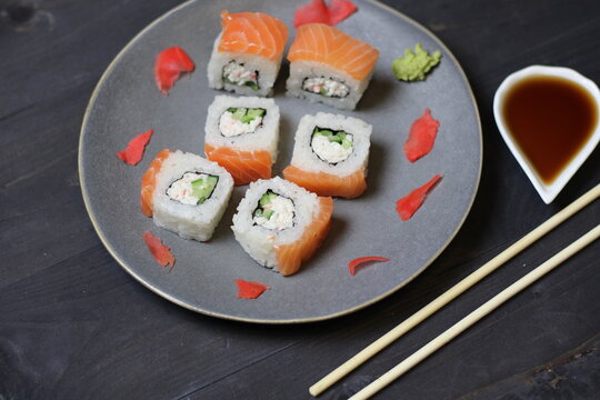 Rolls with salmon, rice, cheese, avocado, caviar, marinated ginger, vasabi on plate with sticks, soy sauce on black background