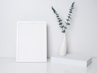 Mock up white wooden poster frame and book decor with  Eucalyptus  leaves in modern ceramic vase  on white table and wall background