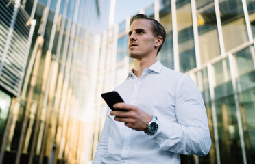 Pensive male entrepreneur 30s waiting for incoming message on cellphone device installing banking app, pondering businessman with mobile technology in hand thoughtful looking away and thinking