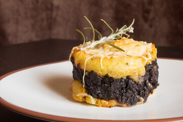 Close-up of shiny chocolate black pudding on a creamy potato sandwich with grated cheese and a fresh sprig of rosemary on top of a wooden table. Homemade recipes food.