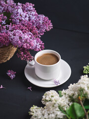 Obraz na płótnie Canvas white cup of fragrant coffee on a dark concrete table, bouquets of white and purple lilacs