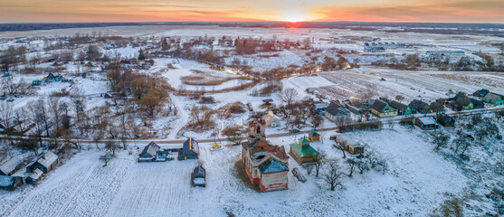 Aerial panorama of old church ruins and modern wooden chapel in the snow at dusk on sunset background. Religion concept. Countryside in winter.