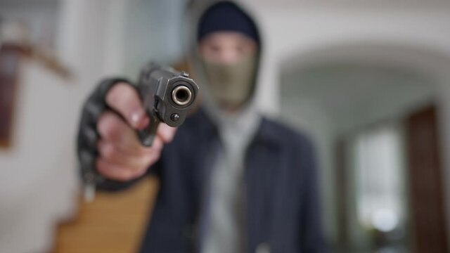 Close-up of gun muzzle with blurred masked man at background aiming. Unrecognizable Caucasian burglar thief threatening wealthy house owner with weapon. Stealing and robbing concept.