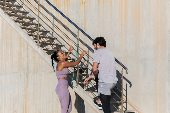 Side view of young sportsman with crossed legs speaking with smiling female partner in sportswear while looking at each other near staircase