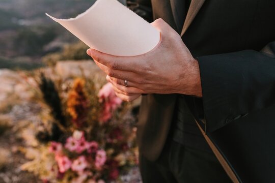 Unrecognizable fiance wearing black suit standing with paper during wedding vow in nature against colorful bouquets of flowers on blurred background